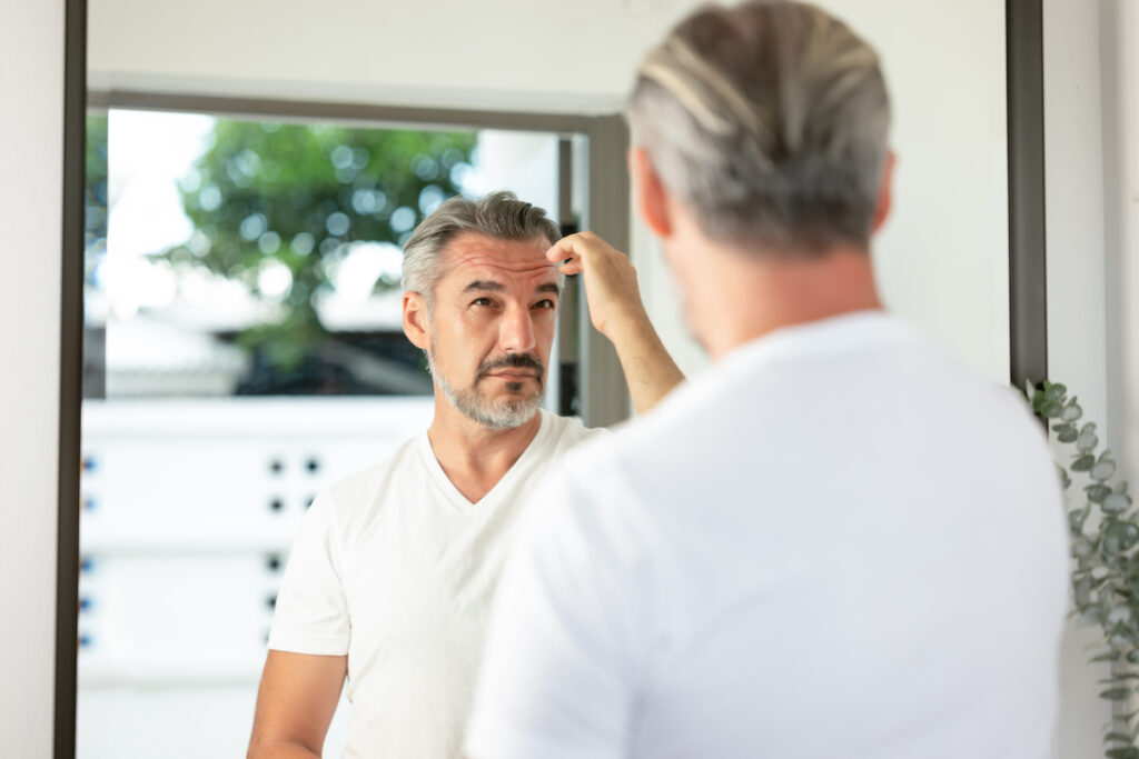 Handsome Caucasian grey-haired man with beard looks in the mirror at his hair after having hair loss treatment in Lawrence