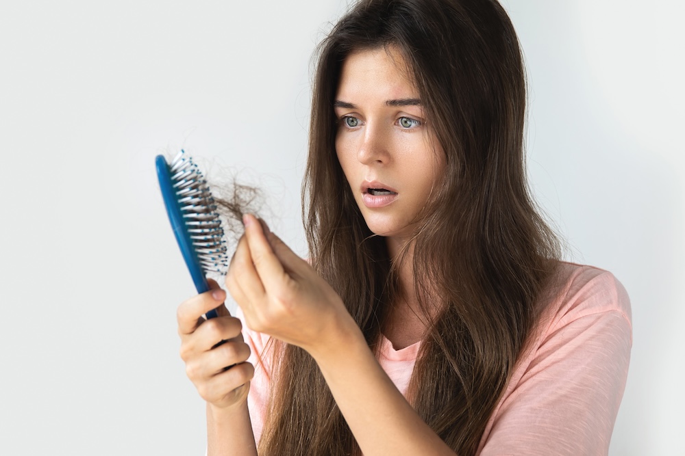 Young woman looks alarmed while pulling a large amount of hair out of her brush, alerting her that it's time to seek hair loss treatment in Leawood