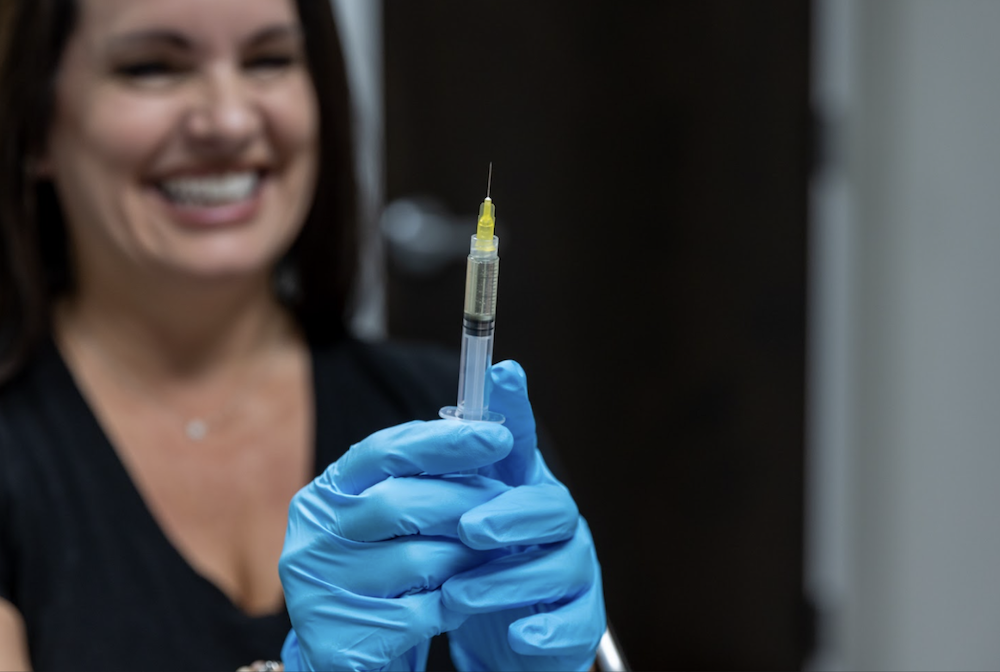 Focused photo of syringe and blurry background of woman holding it and wearing blue gloves to inject bioidentical hormone replacement therapy in Leawood