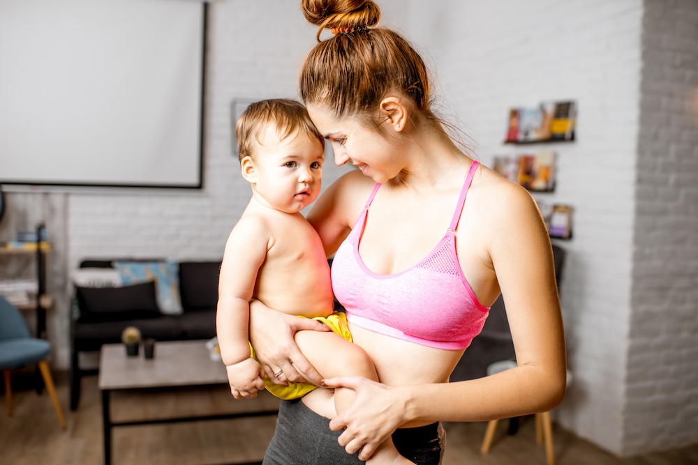 Mother holds baby and wears sports bra after receiving treatment for microneedling for stretch marks in Lawrence.