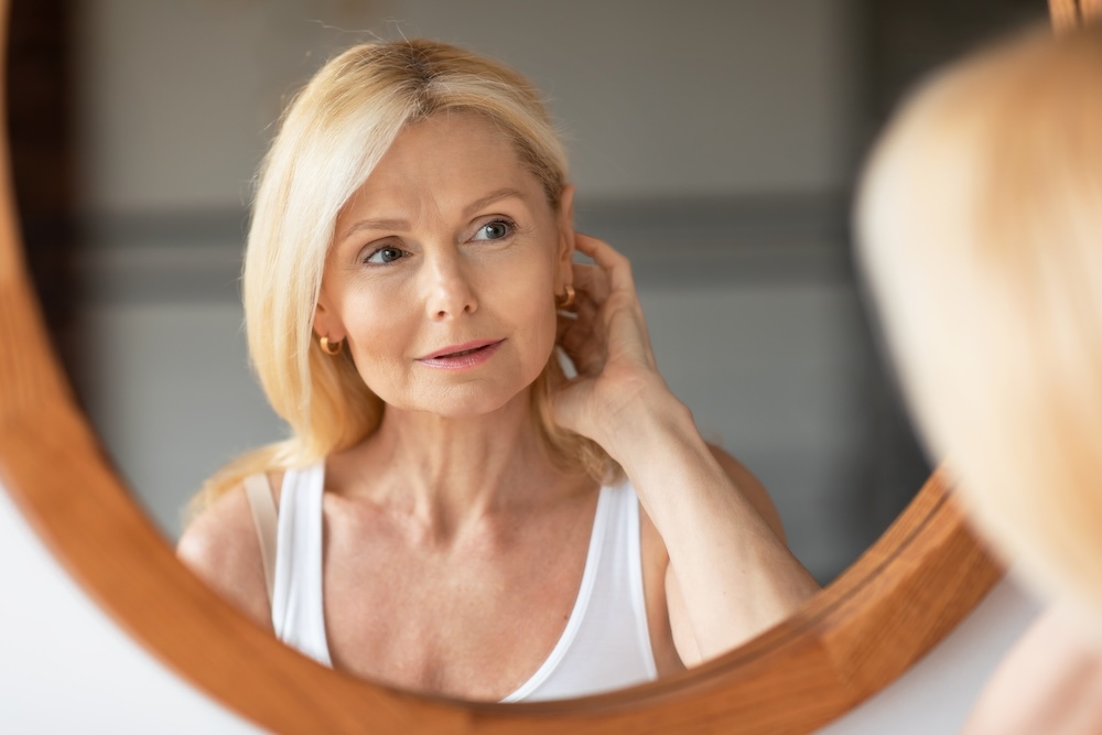 Older woman looks in mirror with hand behind her ear, admiring her reflection because she now has clearer, smoother, tighter skin after getting an injection for Glutathione in Leawood