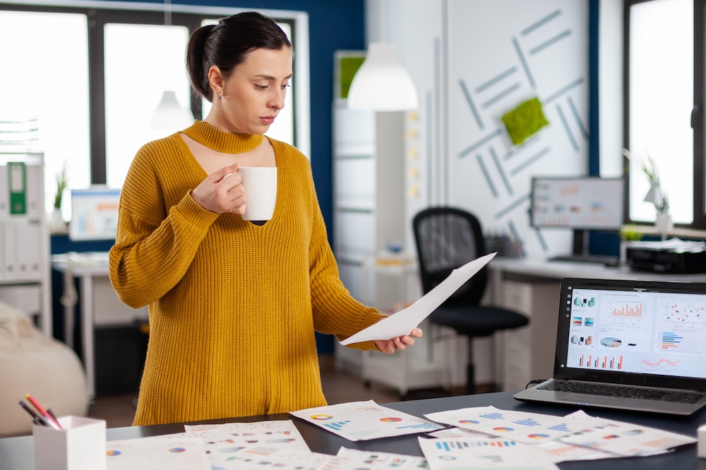 Woman holds coffee cup and stands over a desk with a lap top and papers strewn across her desk as she holds one and analyzes it with renewed focus and energy after her getting an IV for NAD+ in Leawood