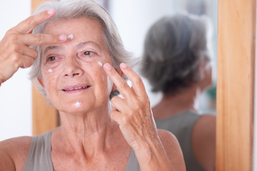 Senior woman applies skin care cream on her forehead, cheeks, and chin as part of a comprehensive plan for age spot removal in Leawood.