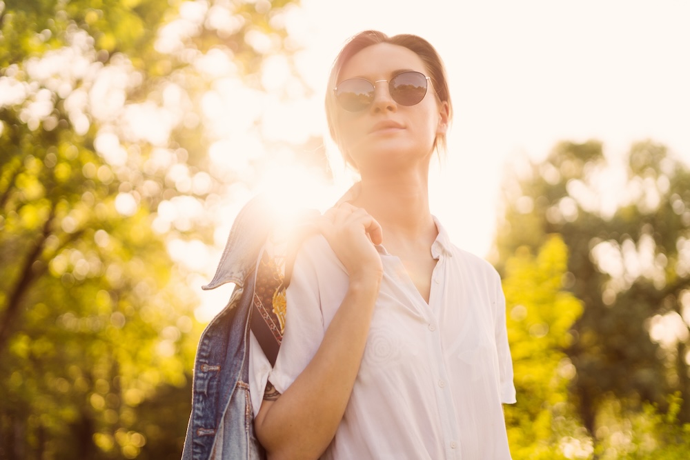 Woman stands in the sunlight, wearing glasses and tosses her denim jacket over her shoulder, unaware that the sun is damaging her skin and she will need laser skin resurfacing in Overland Park.