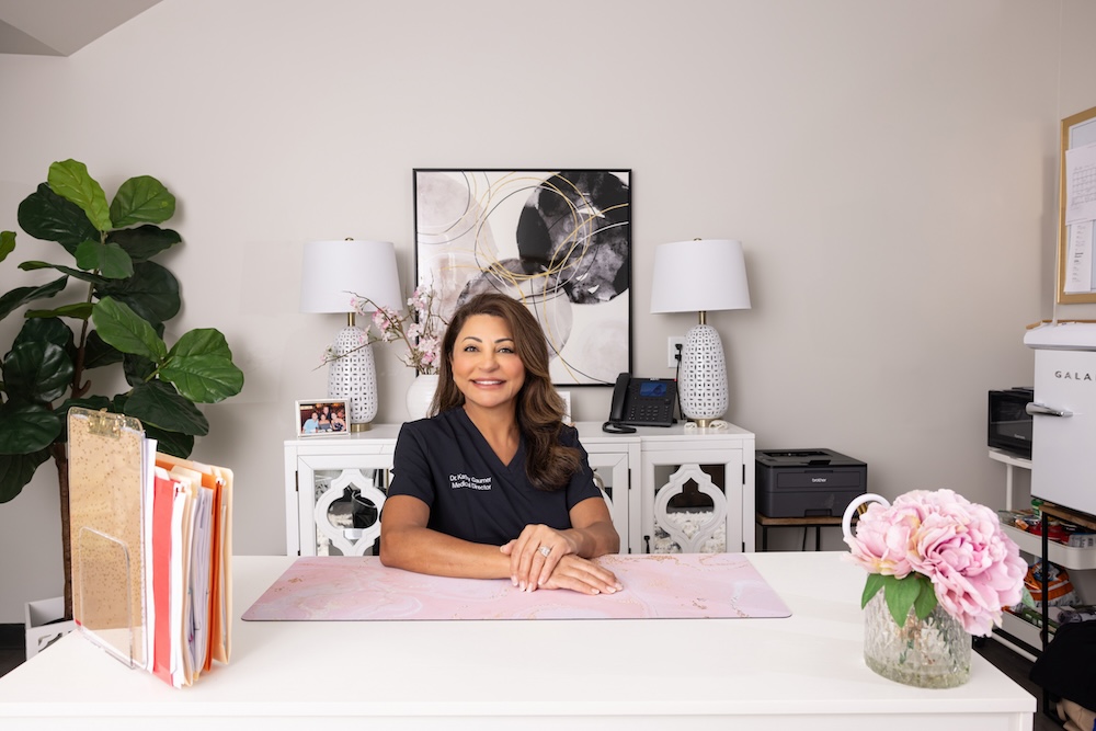 KC med spa owner, Kathy Gaumer, smiles at her desk surrounded by plants, flowers, and decor to make the space feel warm and inviting. On one edge of her desk, you see papers and files placed neatly inside folders and sitting in front of a clipboard to show that she is organized, and ready to meet with clients.
