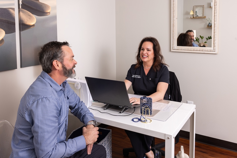 The patient and provider are sitting across from one another at a desk. The patient describes his symptoms and the reason for visiting the wellness center in Kansas City, while the provider takes notes on her laptop to determine the best treatment for him.