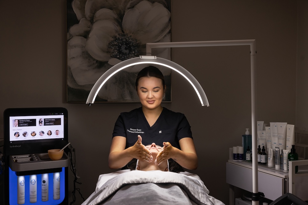 Provider stands over patient who is laying down under a bright light. The provider gently places her hands on the patient's jawline and tilts her head up as part of the treatment for a non-invasive skin rejuvenation near Kansas City.