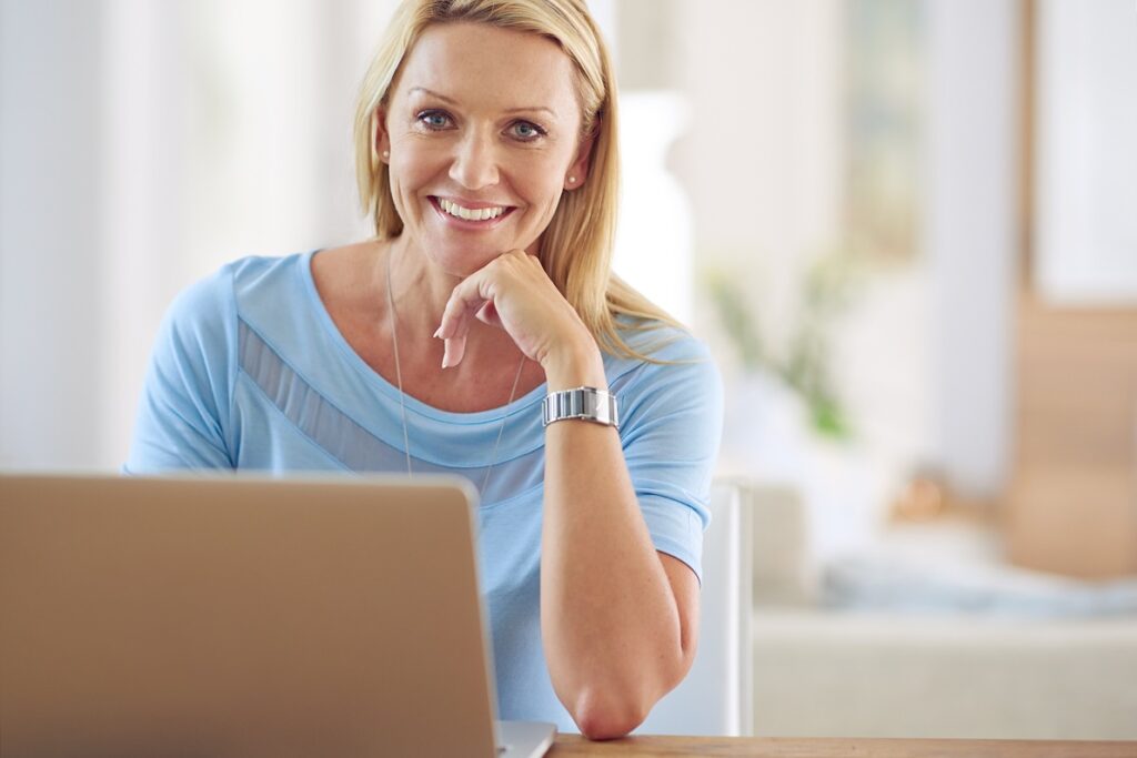Woman smiles with her laptop open. She rests her hand under her chin and gets comfortable for her first consultation with a medical provider through telehealth in Kansas City